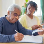 Homem idoso preenchendo documentos enquanto recebe apoio de uma cuidadora familiar em casa, ilustrando o contexto do Carer’s Allowance na Irlanda.