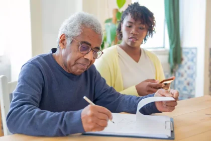 Homem idoso preenchendo documentos enquanto recebe apoio de uma cuidadora familiar em casa, ilustrando o contexto do Carer’s Allowance na Irlanda.