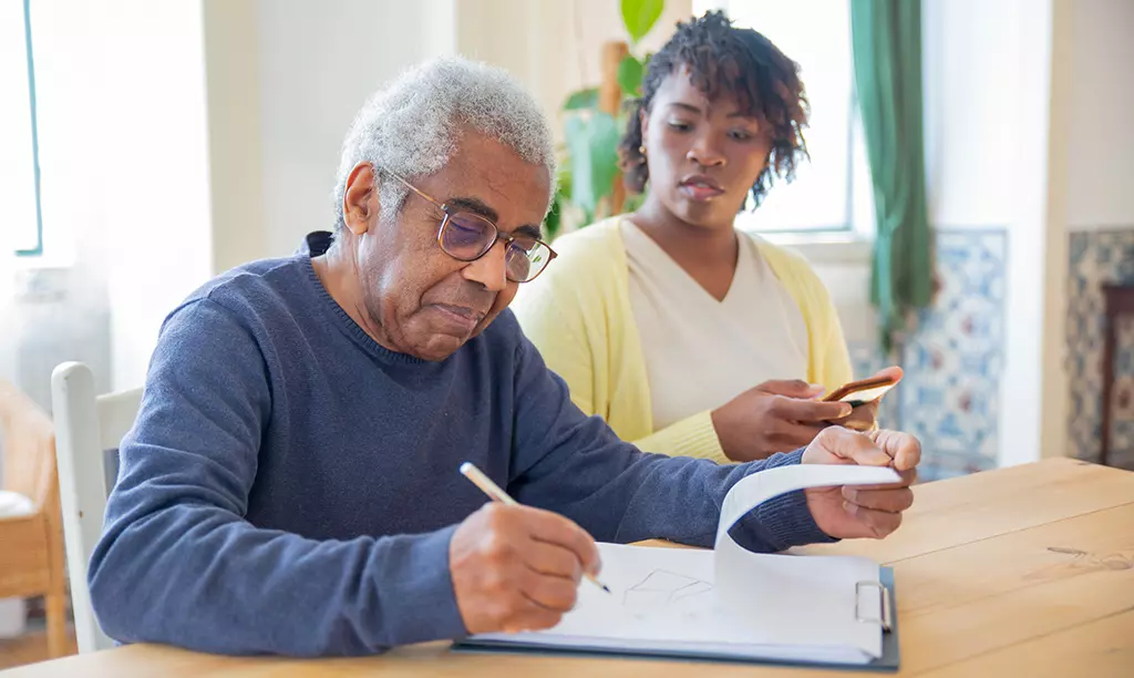 Homem idoso preenchendo documentos enquanto recebe apoio de uma cuidadora familiar em casa, ilustrando o contexto do Carer’s Allowance na Irlanda.
