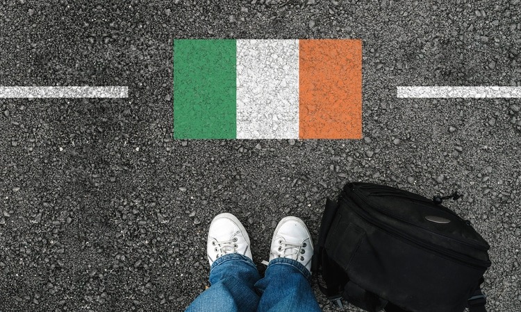Pessoa com mala em frente à bandeira da Irlanda pintada no asfalto, representando imigração e mudança para o país.